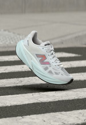 Zapatilla de correr New Balance blanca con suela azul claro, en el aire sobre un paso de peatones de la ciudad con pequeños restos visibles debajo de la zapatilla.