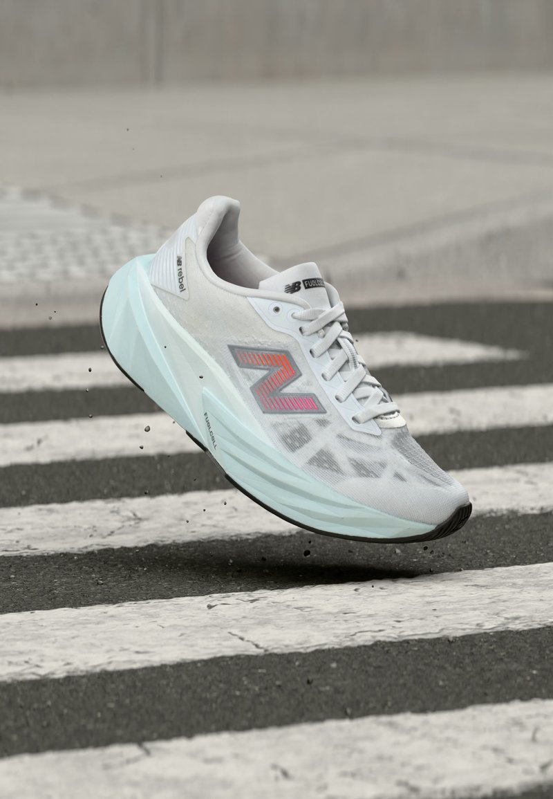 Hvid New Balance løbesko med lyseblå sål, i luften over et fodgængerfelt midt i byen med små synlige skår under skoen.