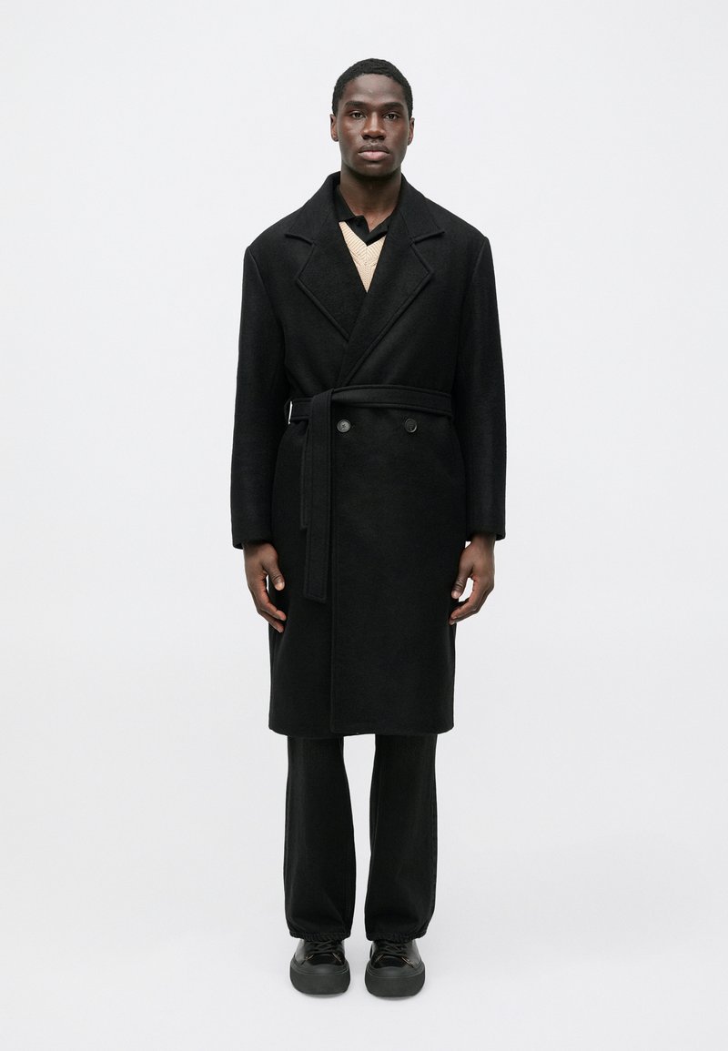 Fekete gyapjúkabát széles hajtókával, kétsoros gombokkal, övvel, bézs kötött pulóver és fekete nadrág fölött viselve, fekete cipővel befejezve.