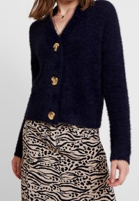 Navy blue fluffy cardigan with a deep V-neck and four large gold buttons, paired with a beige skirt featuring a black animal print pattern.