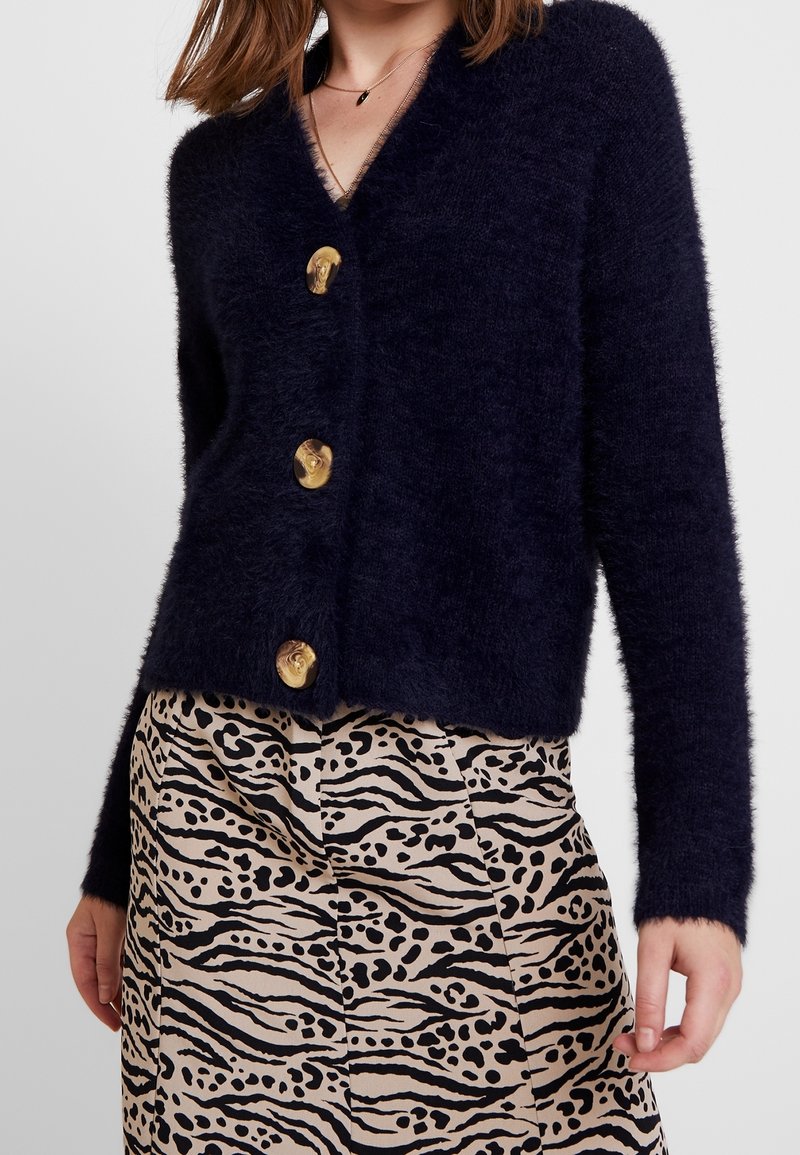 Navy blue fluffy cardigan with a deep V-neck and four large gold buttons, paired with a beige skirt featuring a black animal print pattern.