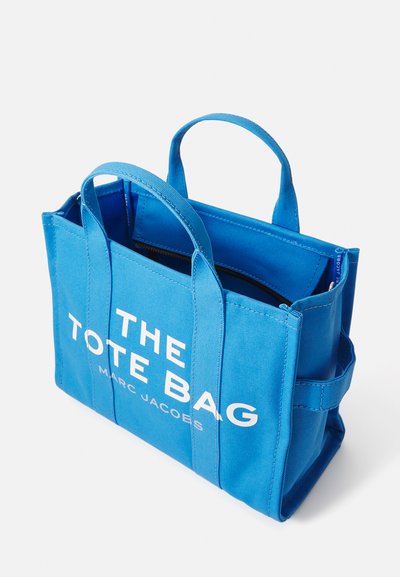 Sac fourre-tout en toile bleue avec double poignées, orné d'inscriptions blanches "THE TOTE BAG" et "MARC JACOBS", forme structurée avec fermeture éclair.