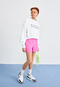 Fehér rövidített pulóver "NIKE" felirattal, rózsaszín sportnadrág, fehér zokni és fekete sportcipő. Egy világoszöld válltáskát tart.