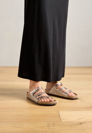 Vaaleat harmaat sandaalit, joissa on kolme säädettävää solkea, korkkipohja ja kumipohja, yhdistettynä mustaan, lattian pituiseen mekkoon. Luonnollinen puulattia.