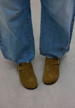 Bruine suède klompen met een ronde neus, twee gespsieraden, gecombineerd met lichtblauwe, wijdvallende jeans, tegen een neutrale achtergrond.