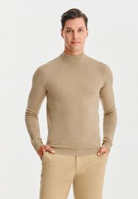 Beżowy sweter z golfem wykonany z miękkiego materiału, o dopasowanym kroju i długich rękawach. Noszony z dopasowanymi beżowymi spodniami.