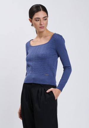 Vrouw met donker haar in een blauwe gestructureerde trui met lange mouwen en zwarte geplooide broek, kijkt naar links met één hand in haar zak.
