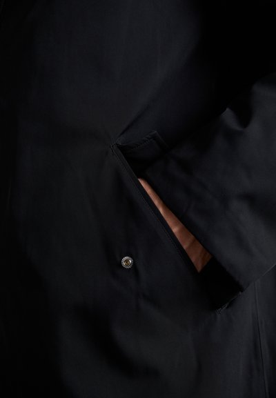 Černý nepromokavý kabát s postranní kapsou na knoflíkové zapínání, hladkou texturou a elegantním designem. Vnitřní podšívka je světlejší barvy.