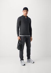 Tamno siva zip-up dukserica s dugim rukavima, uparen s crnim trapericama i srebrnim sportskim cipelama. Mala crna torba se drži u desnoj ruci.