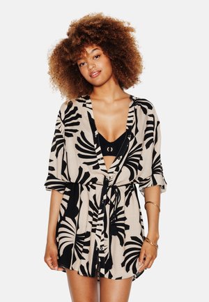 Donna con capelli ricci che indossa un vestito nero e beige con motivo a foglie e bottoni sopra una maglietta nera, accessoriata con braccialetti dorati al braccio destro.