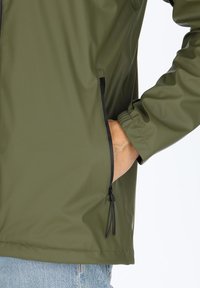 Olivgrüne wasserdichte Jacke mit einer seitlichen Reißverschluss-Tasche und elastischen Bündchen. Glatte Textur mit lässiger Passform und verstellbarem Saum.