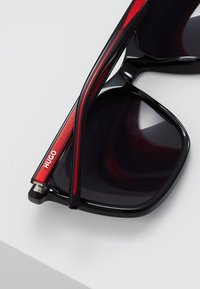 Ochelari de soare negri cu lentile întunecate, accente roșii pe brațele ochelarilor și un detaliu din accesorii argintii. Prezintă un design elegant și modern.