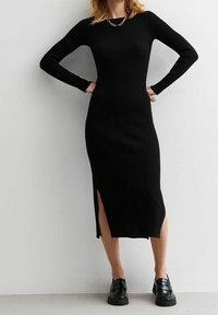 Abito nero a costine con maniche lunghe, nella lunghezza midi e fessure laterali. Caratterizzato da un design aderente e scollatura tonda, abbinato a scarpe nere lucide.