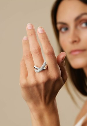 Silberner Ring mit einem verdrehten Design, der eine Reihe kleiner klarer Steine aufweist. Wird an einer Hand mit heller Haut vor einem neutralen Hintergrund getragen.