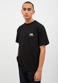 Svart bomullströja med en avslappnad passform, med en färgglad grafisk logotyp på vänster bröst. Korta ärmar och rundad halsringning.