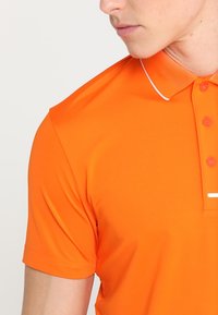 Polo orange en matière synthétique lisse. Comprend un col, trois boutons, et un accent blanc le long du bord du col.