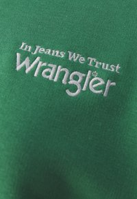 Roheline puuvillane T-särk, millel on valge tikandiga tekst "In Jeans We Trust Wrangler". Sile tekstuur ja lihtne disain.