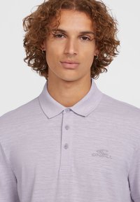 Polo-shirt violet clair avec un col boutonné, en tissu texturé, et un logo brodé sur le côté gauche. La chemise présente une coupe classique.