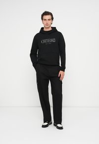 Mężczyzna stojący przy białej ścianie, ubrany w czarną bluzę z kapturem Guess, czarne spodnie cargo i czarno-białe loafersy, z rękami w kieszeniach.