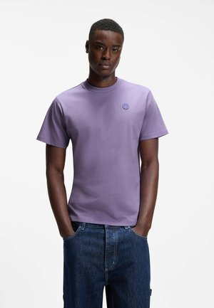 Jeune homme aux cheveux courts portant un t-shirt lavande uni à manches courtes et un jean en denim bleu, debout les mains dans les poches.