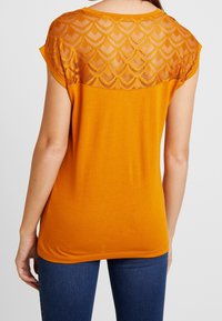 Eine Frau trägt ein kurzärmliges, oranges Oberteil mit einem transparenten Spitzenmuster im Einsatz, kombiniert mit blauen Jeans, von hinten gezeigt.