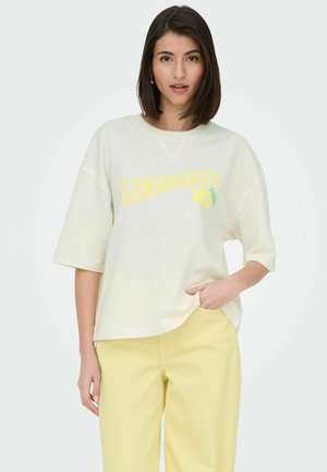 Kobieta z ciemnymi włosami do ramion nosi jasnoszarą, oversize'ową koszulkę z napisem "LIMONATA" i grafiką cytryny, zestawioną z bladożółtymi spodniami.