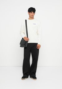 Chemise à manches longues crème avec broderie bleue, pantalon noir à jambes larges et sac bandoulière noir. Modèle portant des espadrilles foncées.