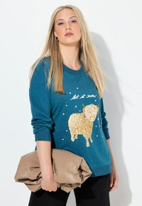 Felpa turchese con una grafica di pecora dorata, testo "let it snow" e motivi di fiocchi di neve bianchi; il modello tiene una pochette beige in una mano.
