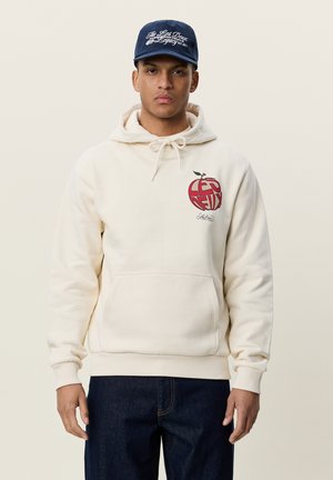 Jeune homme portant un sweat à capuche crème avec un motif de pomme rouge et une casquette bleu marine avec un texte blanc, debout devant un fond clair uni.