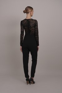 Body en dentelle noire à manches longues, taille cintrée, et pantalon noir fuselé ; le modèle porte des talons hauts noirs. Détails subtils dans le motif de dentelle.