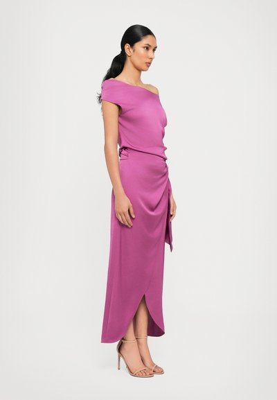 Abito fucsia off-the-shoulder con design drappeggiato e spacco laterale, abbinato a tacchi nudi con cinturini. Tessuto morbido e vestibilità su misura.