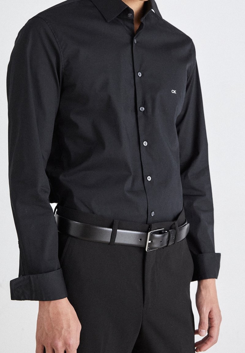 Mies, który ma na sobie czarną koszulę z długim rękawem zapinaną na guziki z logo "CK" oraz czarne spodnie trzymane na skórzanym czarnym pasku.