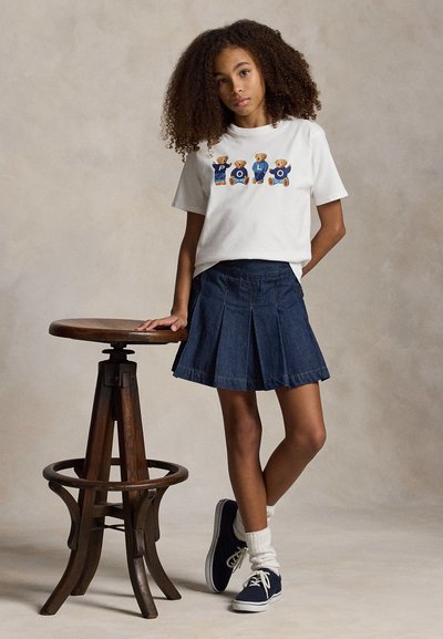 Κορίτσι με σγουρά μαλλιά, που φοράει λευκό μπλουζάκι «POLO» με θέμα αρκουδάκι, μπλε πιετέ φούστα, λευκές κάλτσες και μπλε σκούρα αθλητικά παπούτσια, στηριζόμενο σε ένα ξύλινο σκαμπό.