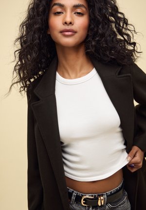Blazer vert foncé sur une chemise blanche ajustée, associé à un jean bleu clair. Boucle de ceinture dorée remarquable et cheveux texturés.