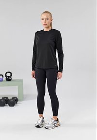 Žena u crnoj sportskoj odjeći stoji u teretani s kettlebellovima i bučicama u blizini na svijetloj pozadini.