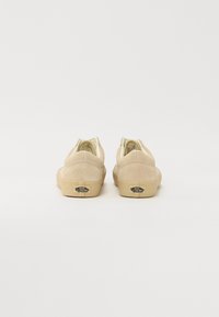 Beige canvas slip-on kengät, joissa on teksturoidun pinnan, kumipohjat ja logovihje kantaosassa, matalaprofiilinen muotoilu.