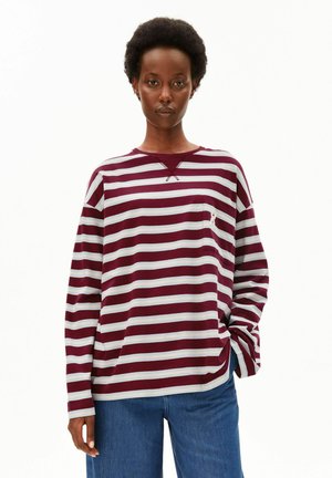 Person trägt ein langärmliges, bordeaux-weiß horizontal gestreiftes Shirt mit kleiner Tasche, kombiniert mit blauen Jeans, steht mit einer Hand in der Tasche.