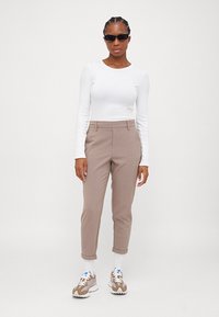 Vit långärmad topp, beige skräddarsydda byxor och ljusbruna sneakers med vita detaljer; modell som bär svarta solglasögon.