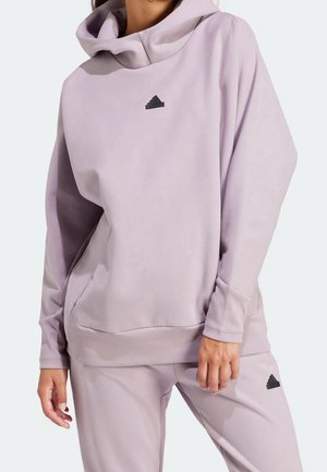 Femme portant un sweat à capuche lilas et un pantalon assorti avec des logos Adidas noirs, mains détendues le long du corps.