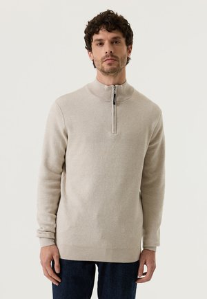 Mann mit kurzem, lockigem Haar trägt einen beigefarbenen Pullover mit Viertelreißverschluss und dunkle Jeans, steht vor einem schlichten, hellen Hintergrund und schaut nach vorne.