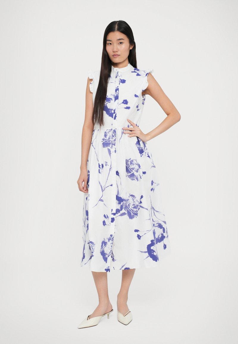 White floral dress with blue patterns, short ruffled sleeves, button front, and a flowy ankle-length skirt. Model wears off-white mules.