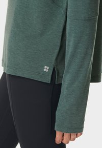 Een persoon in een groen shirt met lange mouwen en een zijsplit, met zwarte leggings, hand ontspannen langs het lichaam tegen een effen achtergrond.