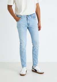 Light blue denim jeans with a slim fit, featuring front pockets, a zip fly, and a button closure. Worn with white sneakers.