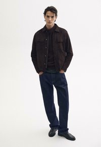 Braune Wildlederjacke mit zwei Brusttaschen, getragen über einem dunklen Oberteil, kombiniert mit dunkelblauen Denim-Jeans und schwarzen Schuhen. Einfacher, lässiger Look.