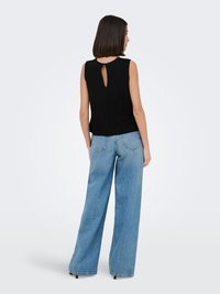 Woman with short dark hair wearing a sleeveless black top with a back keyhole detail and wide-leg light blue jeans standing against a plain background.