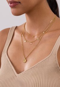 Gestapelte Goldketten mit feinen Ketten, runden Anhängern und einem detaillierten Anhänger, die über einem gerippten beigen Oberteil getragen werden.
