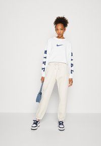 Biela dlhá rukávová tričko Nike s námorníckym logom a nápismi, skombinované s krémovými teplákmi. Doplnky zahŕňajú modrú džínsovú tašku cez rameno a tenisky.