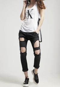 Bílé tílko s černým logem "CK", opotřebované černé džíny s velkými dírami na kolenou a černé šněrovací boty na světlém pozadí.