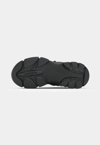 Suela de zapato deportivo negra con un patrón texturizado y un logo prominente. Su diseño incluye múltiples crestas y secciones que mejoran la tracción.