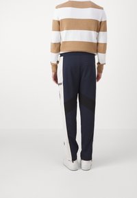 En person klädd i marinblåa och vita träningsbyxor med svarta chevrons och vita sneakers, stående vänd bort från kameran mot en vit bakgrund.